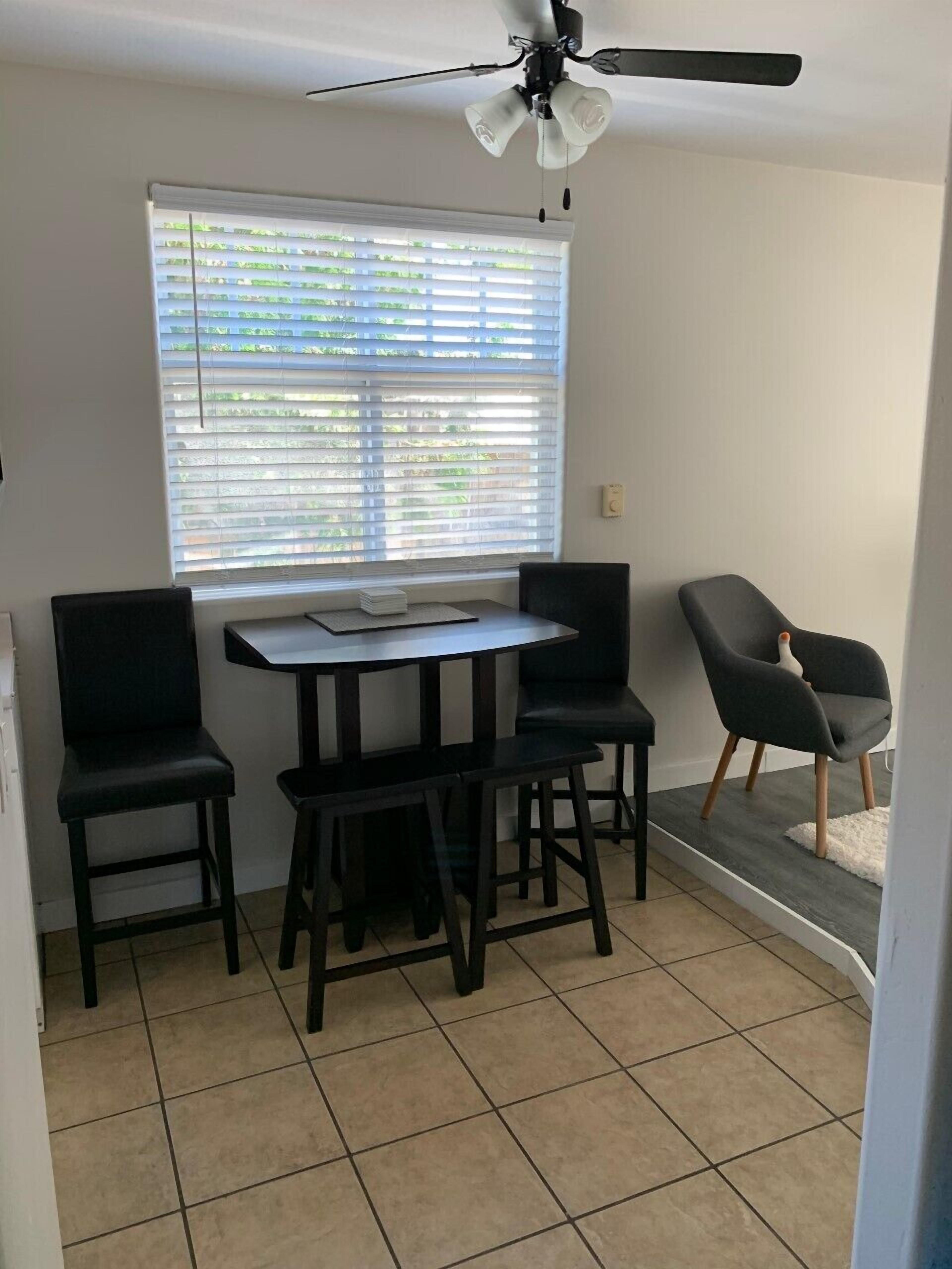 A dining area with a table and four chairs, a ceiling fan, a window with blinds, and a tiled floor.