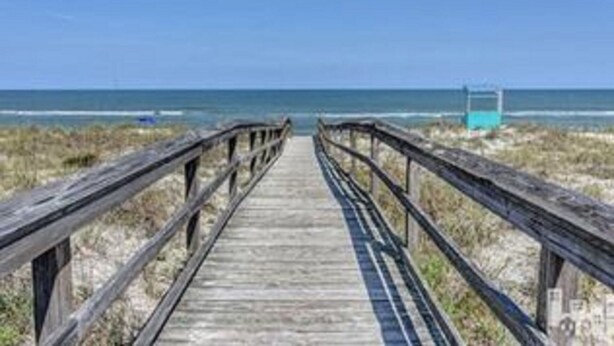 Un muelle de madera que lleva a una playa, con una torre de salvavidas a lo lejos.