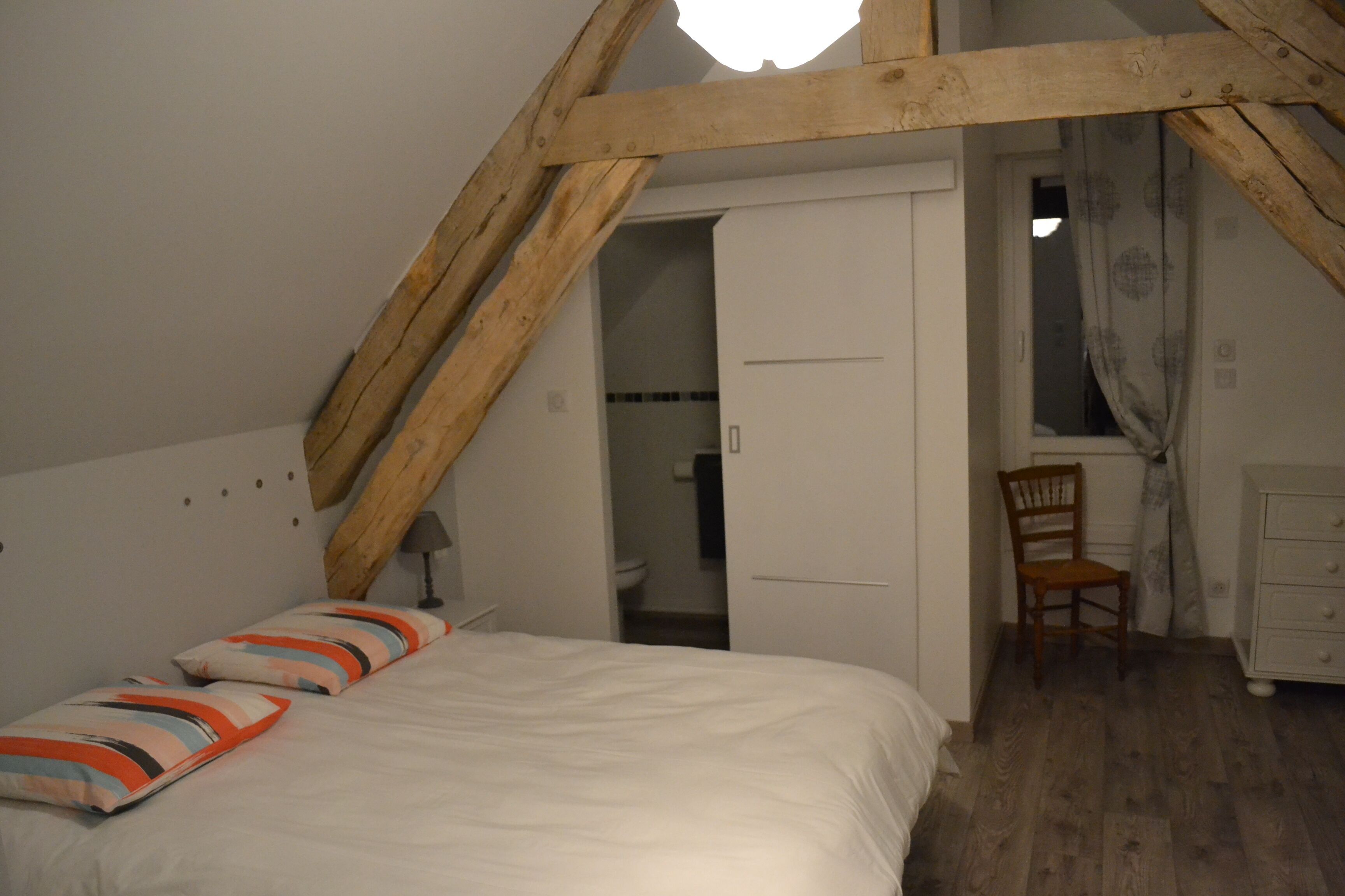 Une chambre avec un plafond en pente, un lit avec des coussins à rayures, une chaise en bois et une commode.