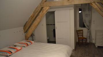 Een slaapkamer met een schuine kap, een bed met gestreepte kussens, een houten stoel en een dressoir.