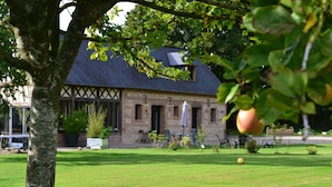 Ein traditionelles Backsteingebäude mit einem geneigten Dach und einem sichtbaren Schornstein. Vor dem Gebäude erstreckt sich eine gepflegte Rasenfläche mit vereinzelten Stühlen und einem Tisch.