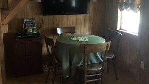 A wooden dining area with a table covered in a green tablecloth, wooden chairs, a television mounted on the wall, and a window with curtains. - Lakeside Cottage, Fishing, Canoe & Kayak, Pet Friendly, Golf and Hiking Nearby (Crofton)