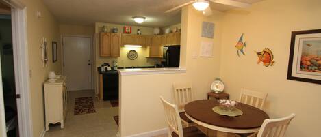 A dining area with a round table and four chairs, a ceiling fan, and a kitchen in the background.