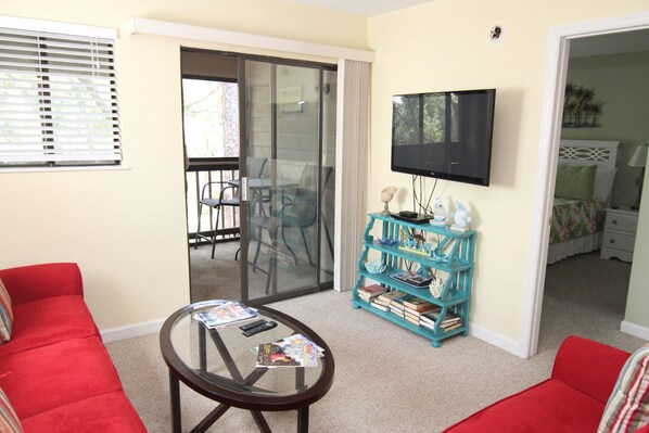 A living room with a red sofa, a glass coffee table, a teal bookshelf, and a TV mounted on the wall.