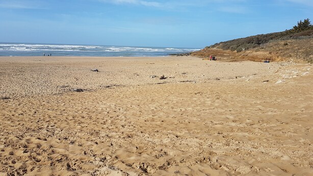 Una extensa playa de arena con olas suaves, cielo despejado y una zona colinosa con vegetación escasa.