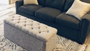 A dark grey sofa with two white cushions, a grey tufted ottoman, and two framed artworks on the wall. - Home away from Home!!  (Boise)