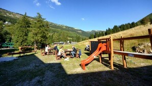 Un parque infantil con un tobogán y mesas de picnic en un área verde, con montañas al fondo.