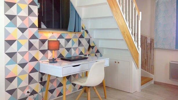 A modern room with a white desk, a lamp, and a chair. There's a window with blinds and a geometric-patterned wall. A wooden staircase is visible in the background. - Triplex, Beautiful apartment of 50 sqm on 3 levels, up to 4 people (Caen)