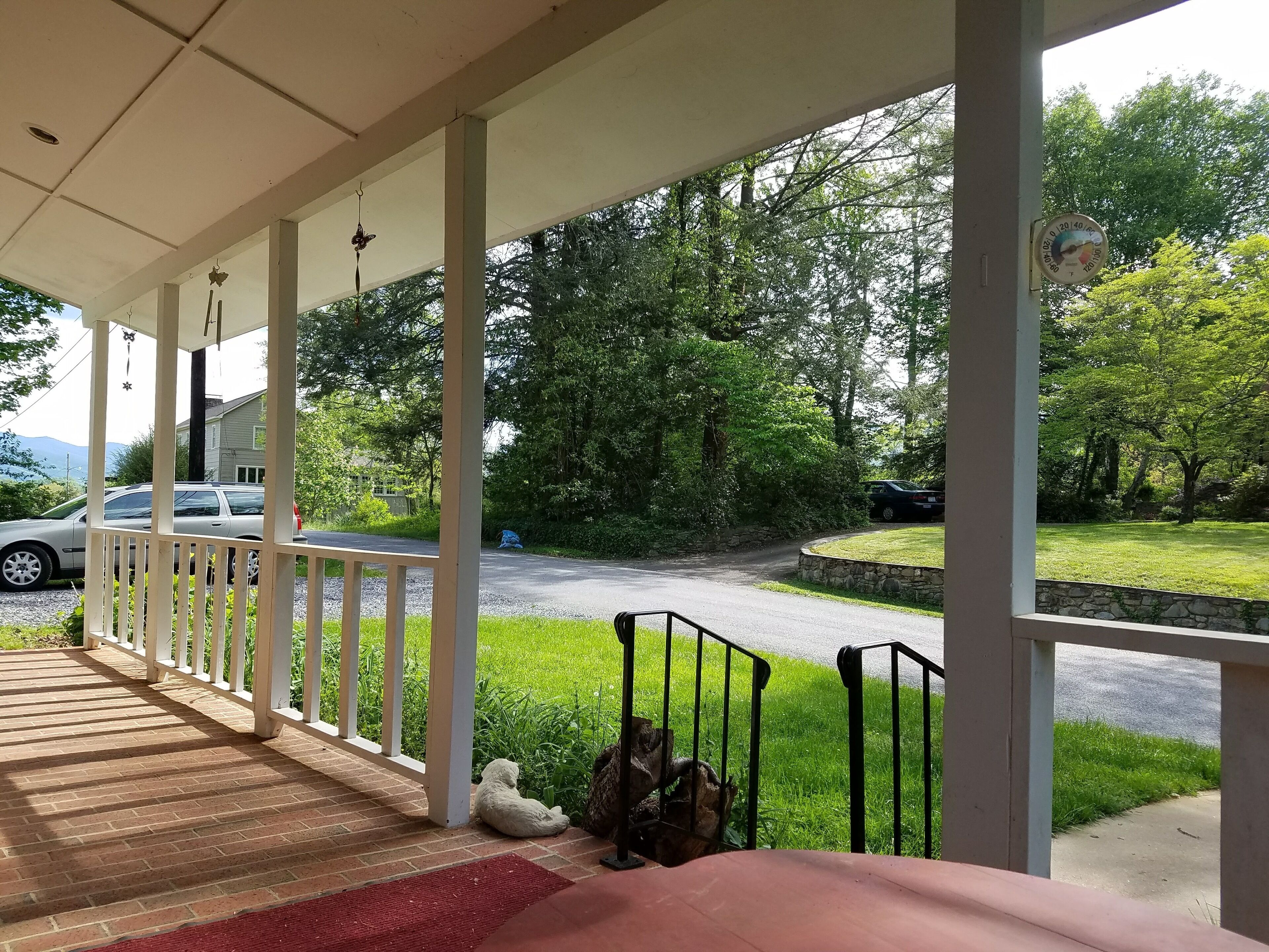 A covered porch with a brick floor and white railings, overlooking a grassy yard and a road with parked cars.