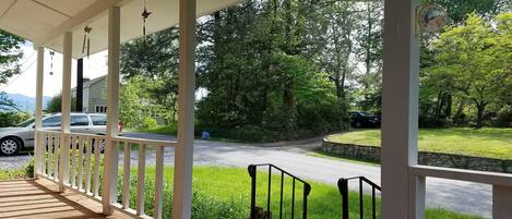 A covered porch with a brick floor and white railings, overlooking a grassy yard and a road with parked cars.