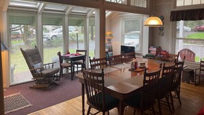 A spacious dining area with a large wooden table and multiple chairs, large windows with blinds, and a view of the outside.