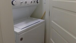 A compact laundry room with a front-loading washer and dryer, a small shelf, and a door leading to another room.