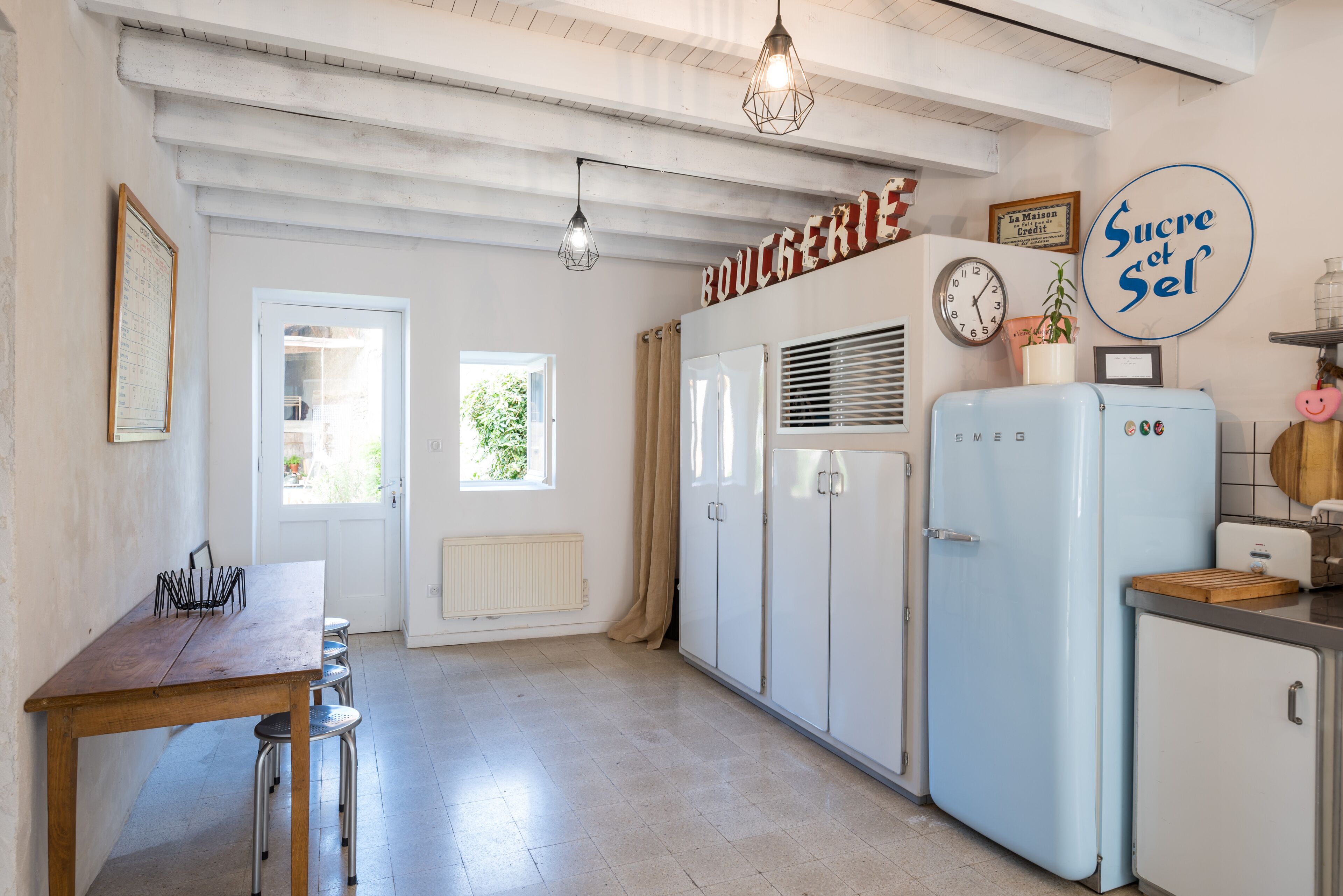 Una cocina con una mesa de madera, armarios blancos, un refrigerador azul y un reloj en la pared.