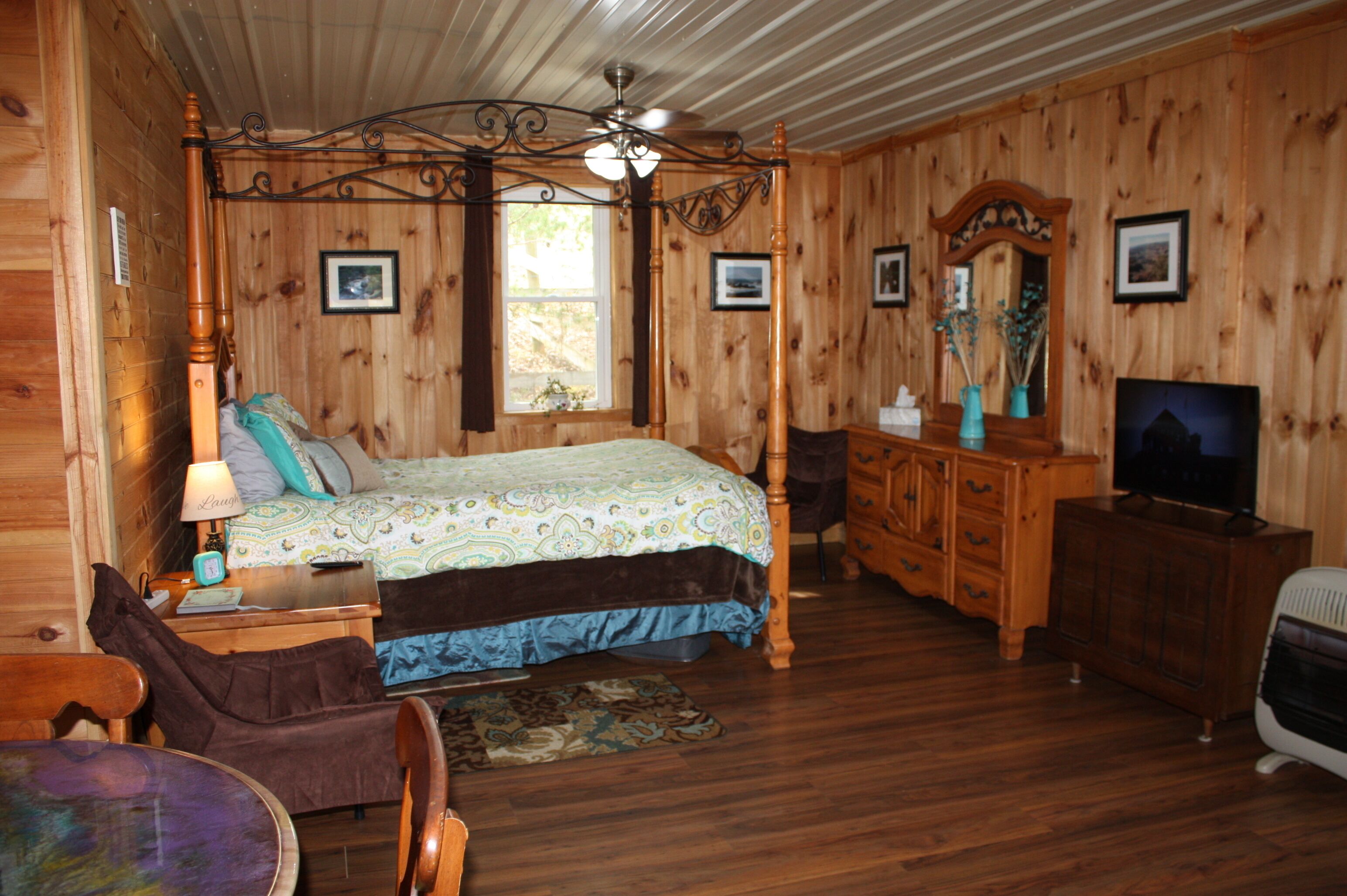 A wooden cabin bedroom with a four-poster bed, a dresser, a television, and a desk with a lamp.