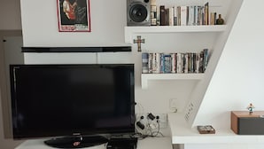 Ein Wohnzimmer mit einem Flachbildfernseher, Bücherregalen, einem Lautsprecher und einer kleinen Figur auf einem Tisch.