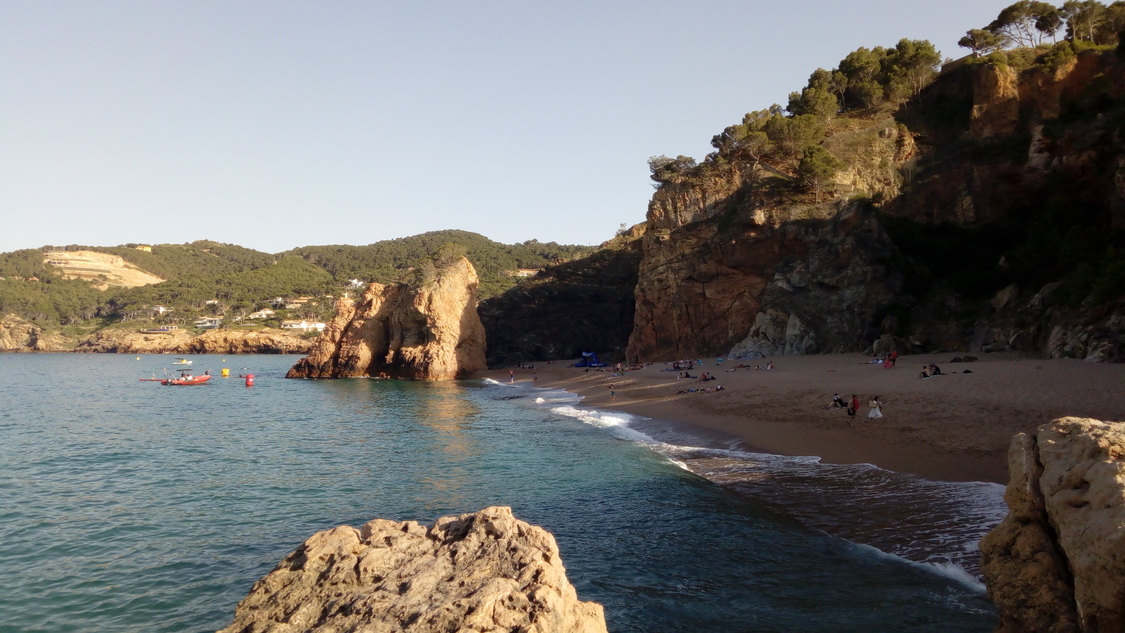 A coastal beach with rocky cliffs, clear blue water, and a few boats.