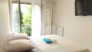 Ein Hotelzimmer mit Bett, einem Vorhangfenster, einem an der Wand befestigten Fernseher und einem Balkon mit grüner Bepflanzung.