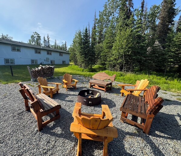 An outdoor seating area with Adirondack chairs and a fire pit, surrounded by gravel and trees.