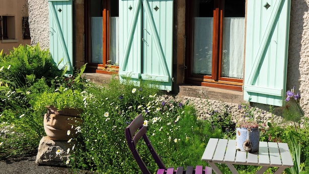 Un bâtiment doté de trois fenêtres, chacune ornée de volets verts. Un jardin, où poussent diverses plantes et fleurs, se trouve devant le bâtiment.