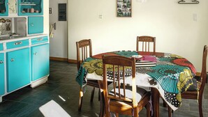 Un coin repas avec une table ronde recouverte d’une nappe colorée, entourée de chaises en bois. On aperçoit à l’arrière-plan une cuisine aux tons turquoise, avec des placards et un évier.