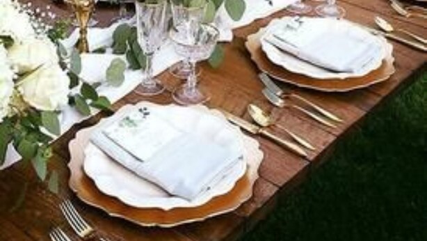 Una larga mesa de madera dispuesta para un evento formal al aire libre, con manteles blancos, platos con detalles dorados y altas velas blancas.