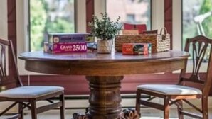 Una mesa redonda de madera con base de león, rodeada de sillas, en una habitación con ventanas grandes y vistas al verdor exterior.