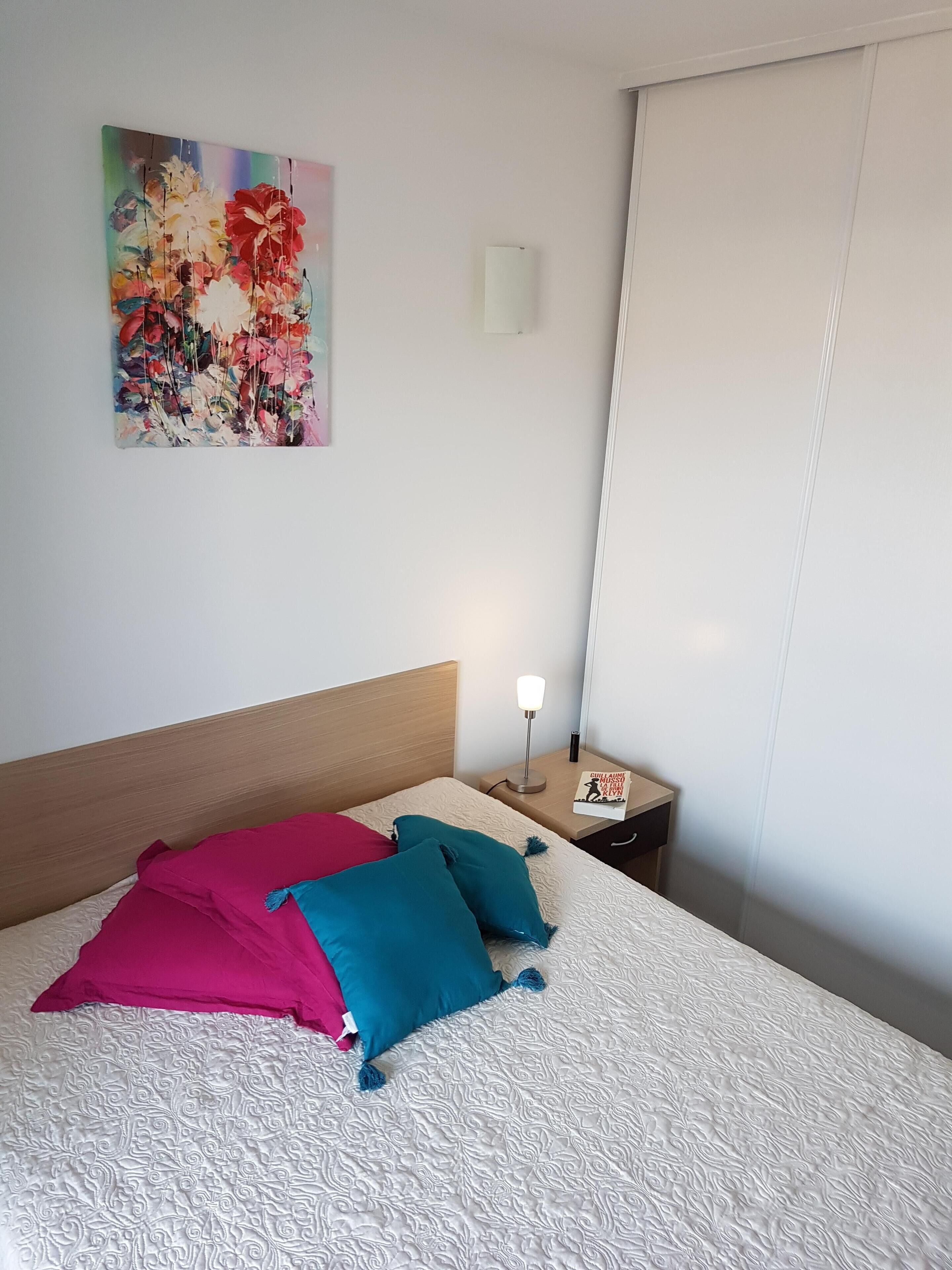 Een net opgemaakt bed met beddengoed met patroon, een kleurrijk schilderij aan de muur, en een nachtkastje met een lamp en een boek.
