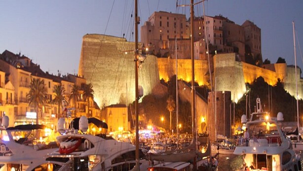 Un porticciolo con barche ormeggiate, un'imponente struttura simile a un castello sullo sfondo e edifici con balconi.