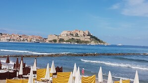 Vista sulla costa con un edificio fortificato storico su una collina, ombrelloni e lettini da spiaggia.