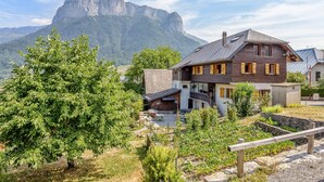 Une maison à deux étages, dotée d’un toit en pente et de volets en bois, entourée d’un jardin avec un grand arbre et un mur en pierre.