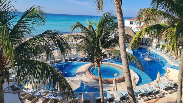 Zwembadgedeelte van een resort met ligbedden en palmbomen, met uitzicht op een strand en zee.