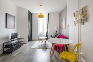 A modern living room with a TV, a small table, colorful chairs, and a hanging light fixture. - Nice Furnished Apartment Close to Amenities (Villeurbanne)