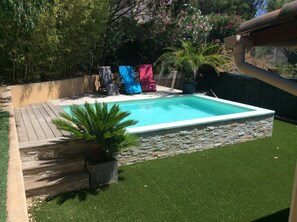 Une piscine rectangulaire bordée de pierres, entourée d’une terrasse en bois et d’une pelouse artificielle.