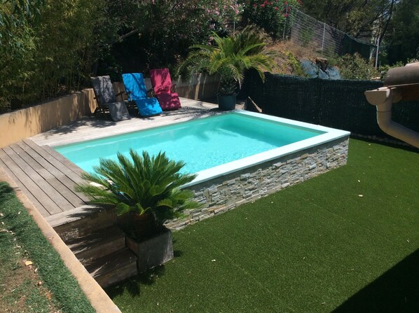 Une piscine rectangulaire bordée de pierres, entourée d’une terrasse en bois et d’une pelouse artificielle.