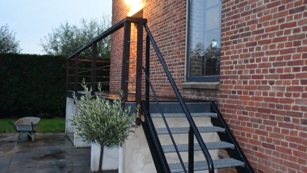 A brick building with a staircase leading to a door, a potted plant, and a wheelbarrow in the foreground.