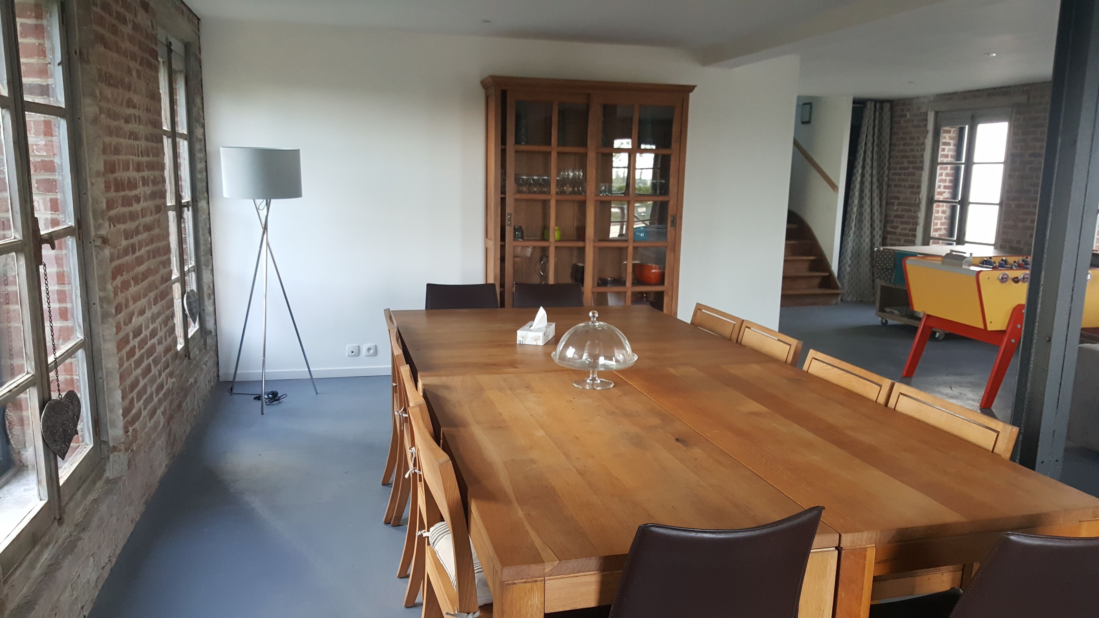 Uma ampla sala de jantar com uma longa mesa de madeira, cadeiras, um centro de mesa em formato de cúpula de vidro, um armário de madeira com portas de vidro, e vista para uma escada e uma mesa de pebolim ao fundo.
