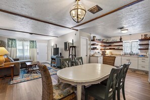 A dining area with a round table and chairs, a television, and a kitchen with shelves and cabinets.