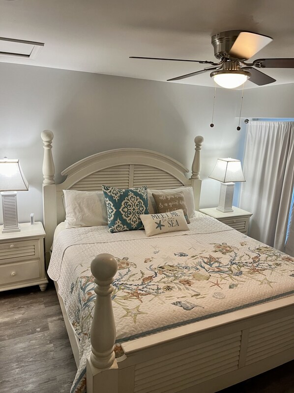 Un dormitorio con cama blanca, almohadas decorativas, ventilador de techo, lámparas de noche y ventana con cortinas.