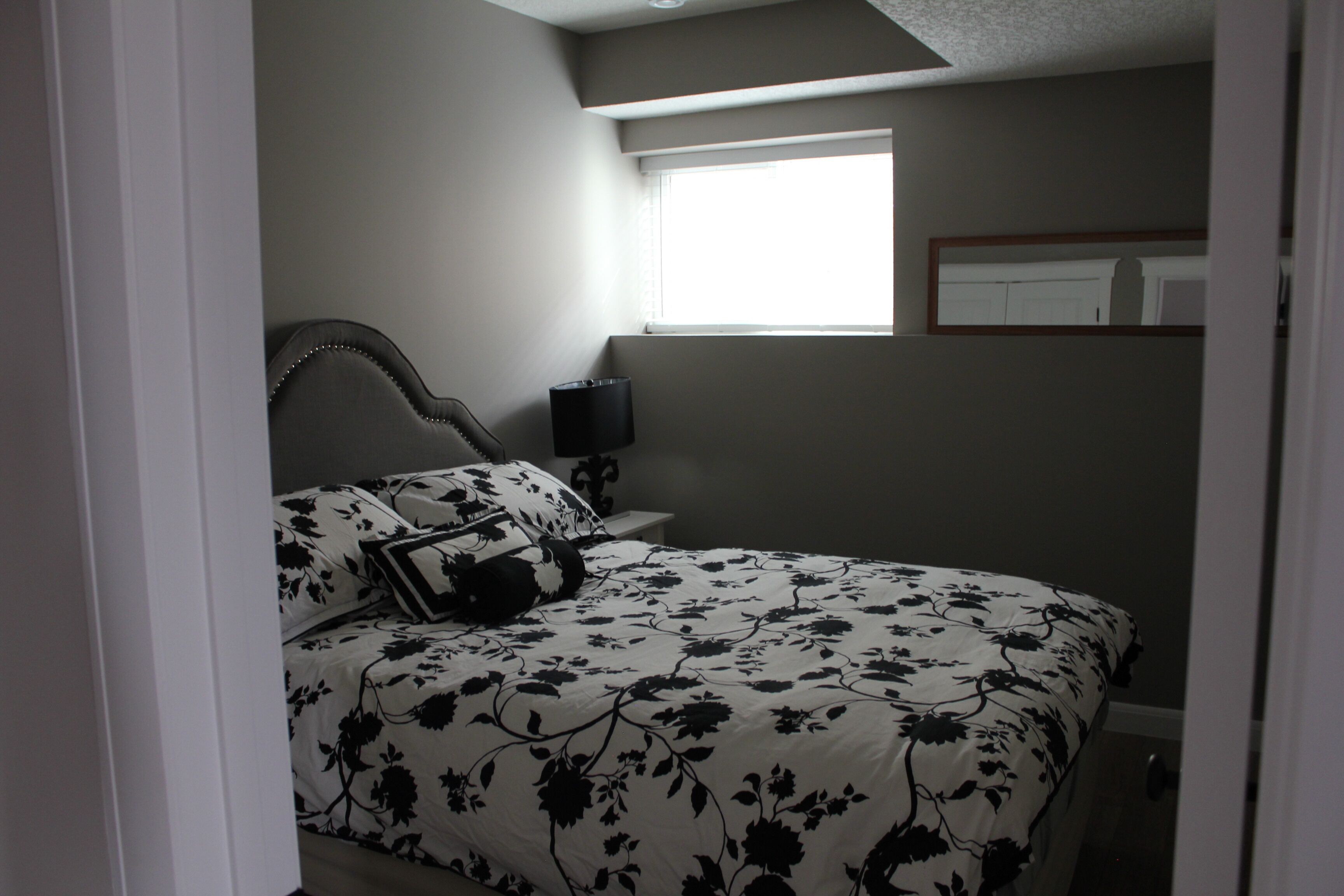 A bedroom with a bed, a nightstand, a lamp, a window, and a mirror on the wall.