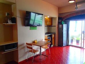 A compact kitchen with a dining table and chairs, a microwave, a refrigerator, and a television mounted on the wall.