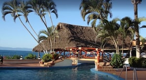 A pool area with a thatched-roof structure, palm trees, and a view of the ocean.