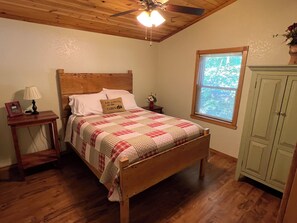 A bedroom with a wooden bed, a nightstand with a lamp, a window with a view, and a green cabinet. - Exceptional Cabin on Shawnee Wine Trail near Alto Pass and Bald Knob Cross (Alto Pass)