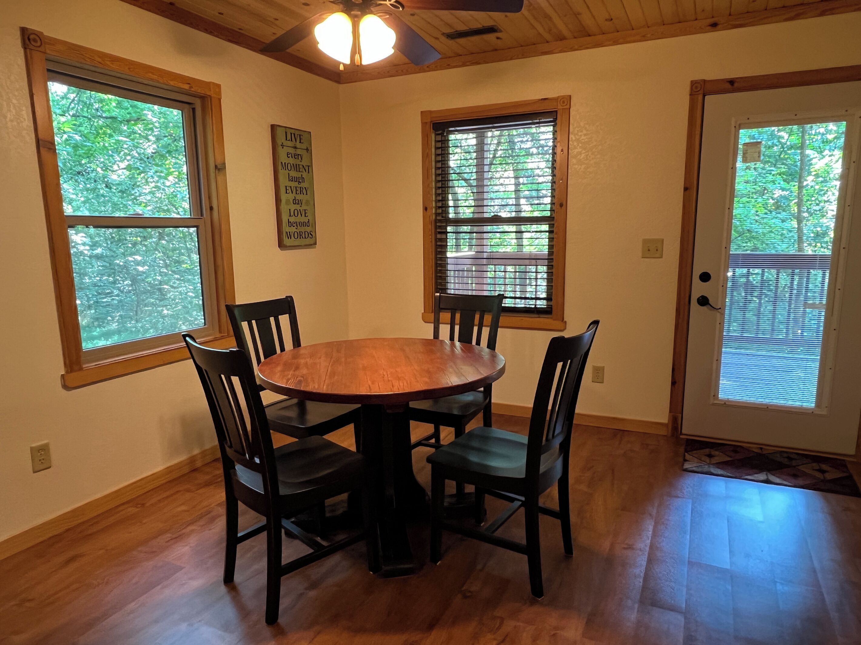 A dining area with a wooden table and chairs, large windows with blinds, and a ceiling fan.