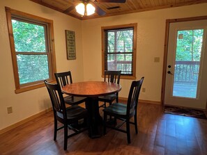 A dining area with a wooden table and chairs, large windows with blinds, and a ceiling fan.