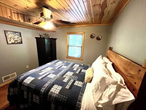 A bedroom with a wooden ceiling, a bed with a quilt, a window with blinds, and a ceiling fan.