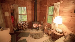 A cozy cabin interior with a wood-burning stove, a sofa, a lamp, and a window overlooking the forest.