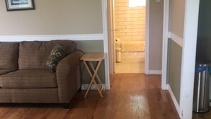 Un salon avec un canapé marron, une petite table en bois et une porte donnant sur une salle de bain dont un mur est recouvert de carreaux blancs.