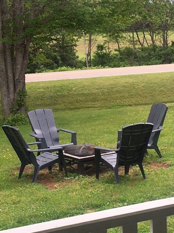 A deck with three Adirondack chairs and a small table, surrounded by a grassy area with trees in the background.