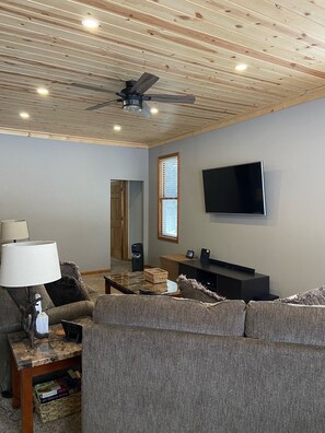 A living room with a ceiling fan, a flat-screen TV mounted on the wall, a sofa, a coffee table, and a lamp. - Your UP Retreat w/ Garage! (Bergland)