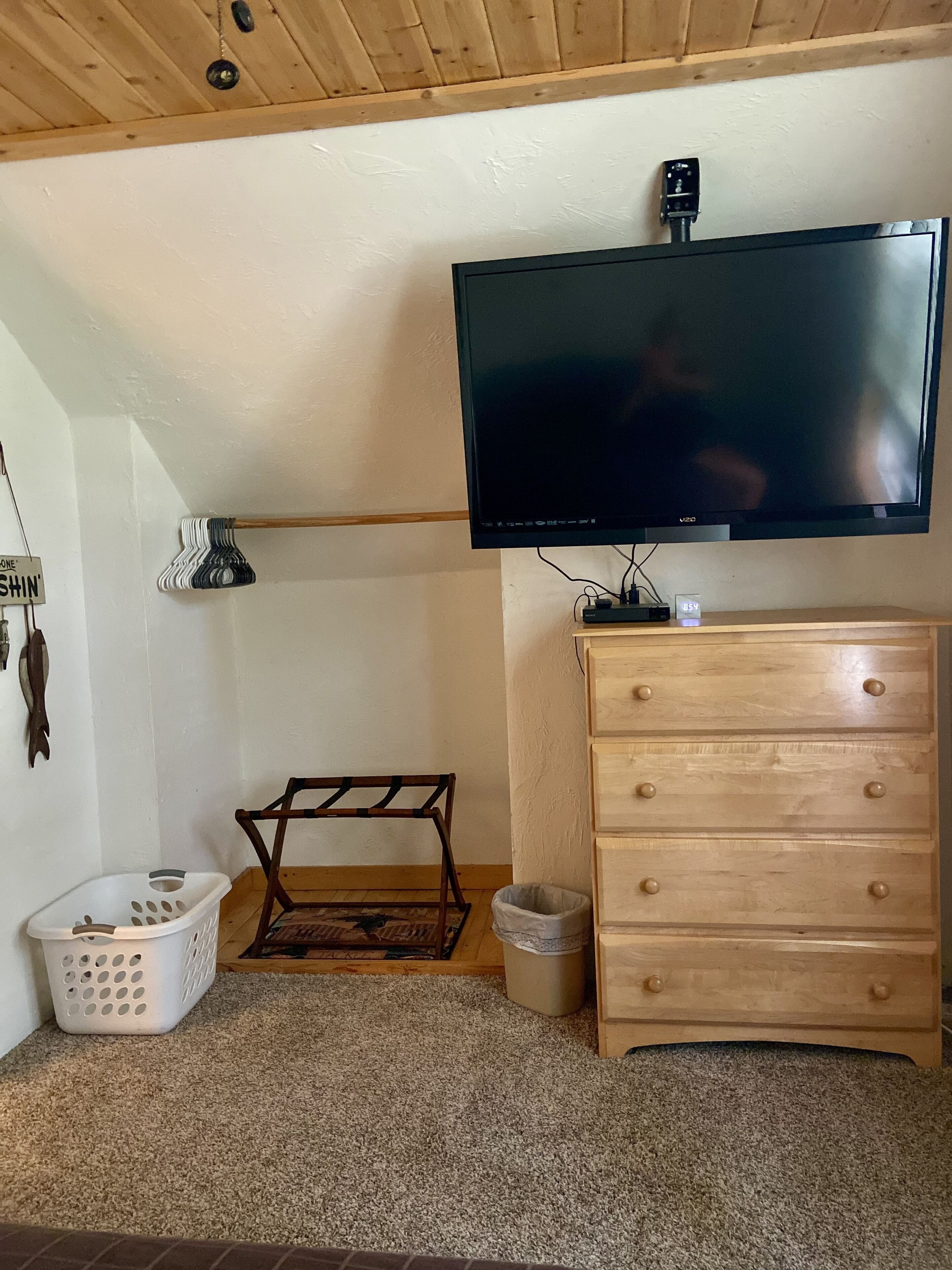 Une pièce avec un téléviseur fixé au mur, une commode en bois, une petite chaise en bois, un panier à linge et une poubelle.